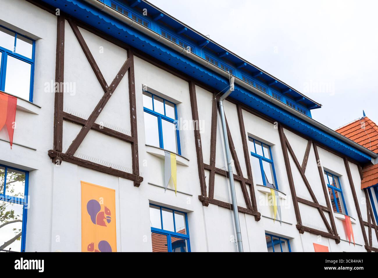 Edificio moderno con finestre incorniciate in blu, design a metà legno, striscioni colorati, vibranti dettagli architettonici sotto il cielo nuvoloso. Foto Stock