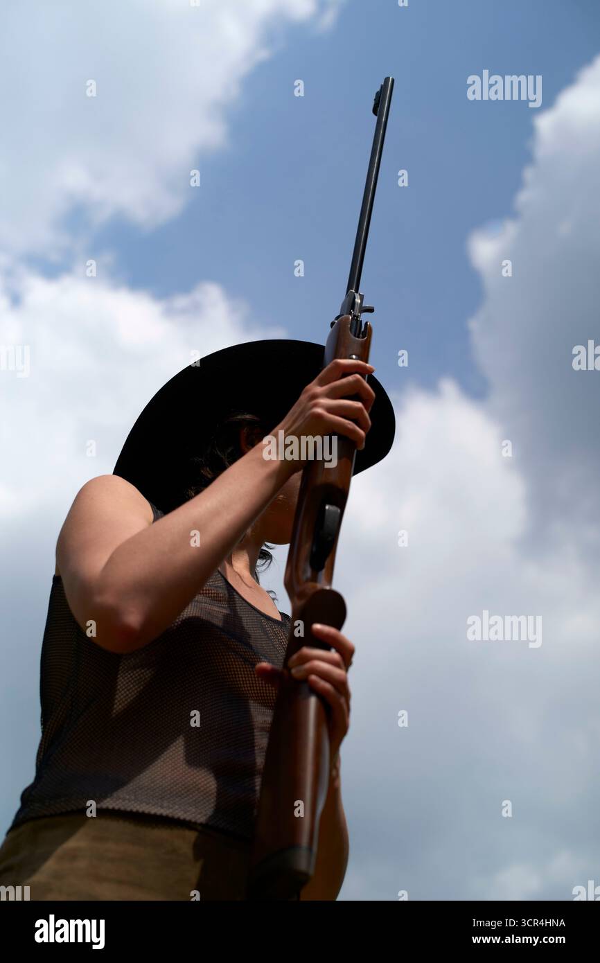 Una donna con un cappello da cowboy regge un fucile su uno sfondo nuvoloso. Foto Stock