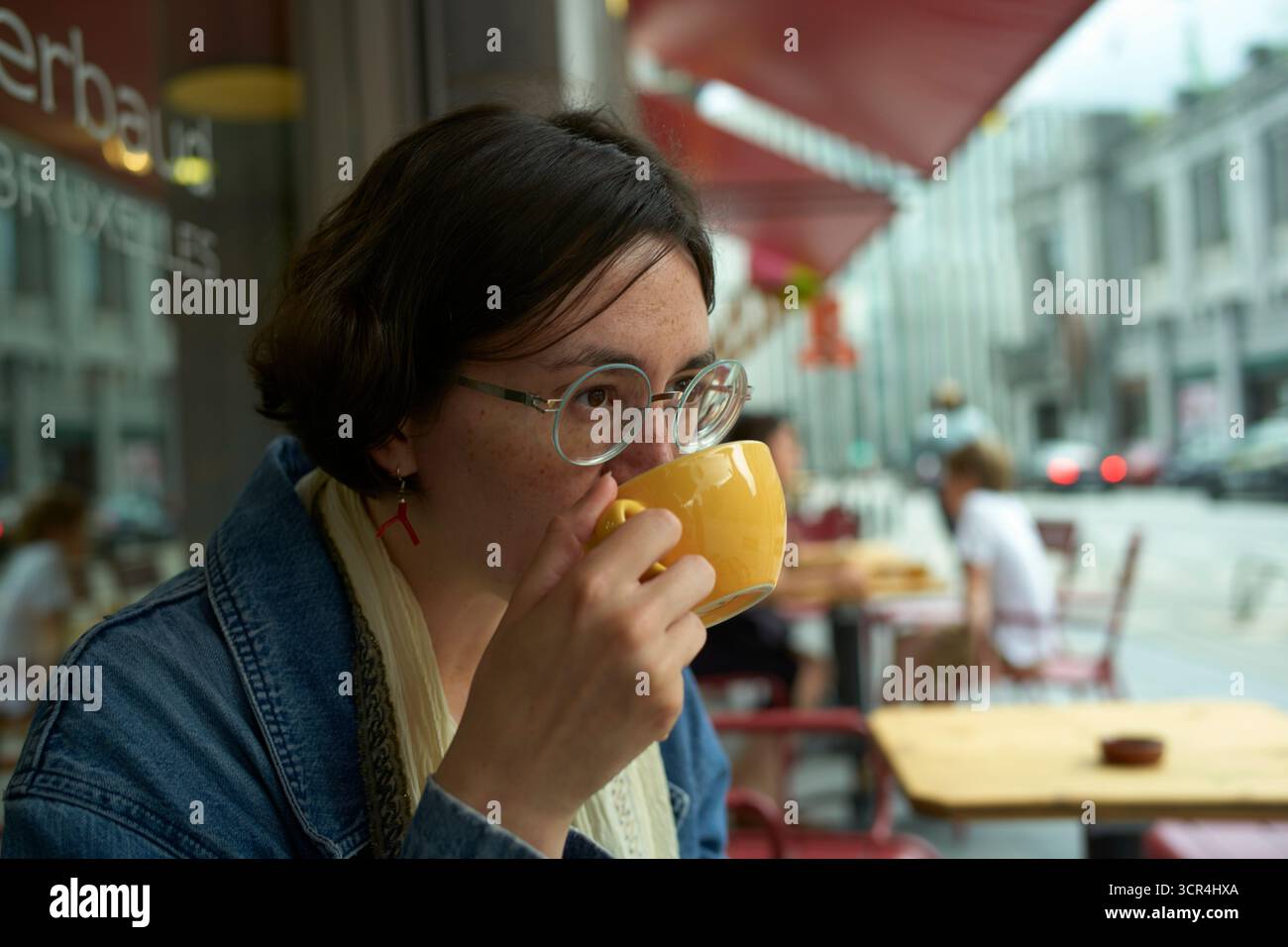 Una donna in occhiali sorseggia un caffè all'aperto in una strada cittadina, indossando una giacca in denim. Bruxelles, Belgio Foto Stock
