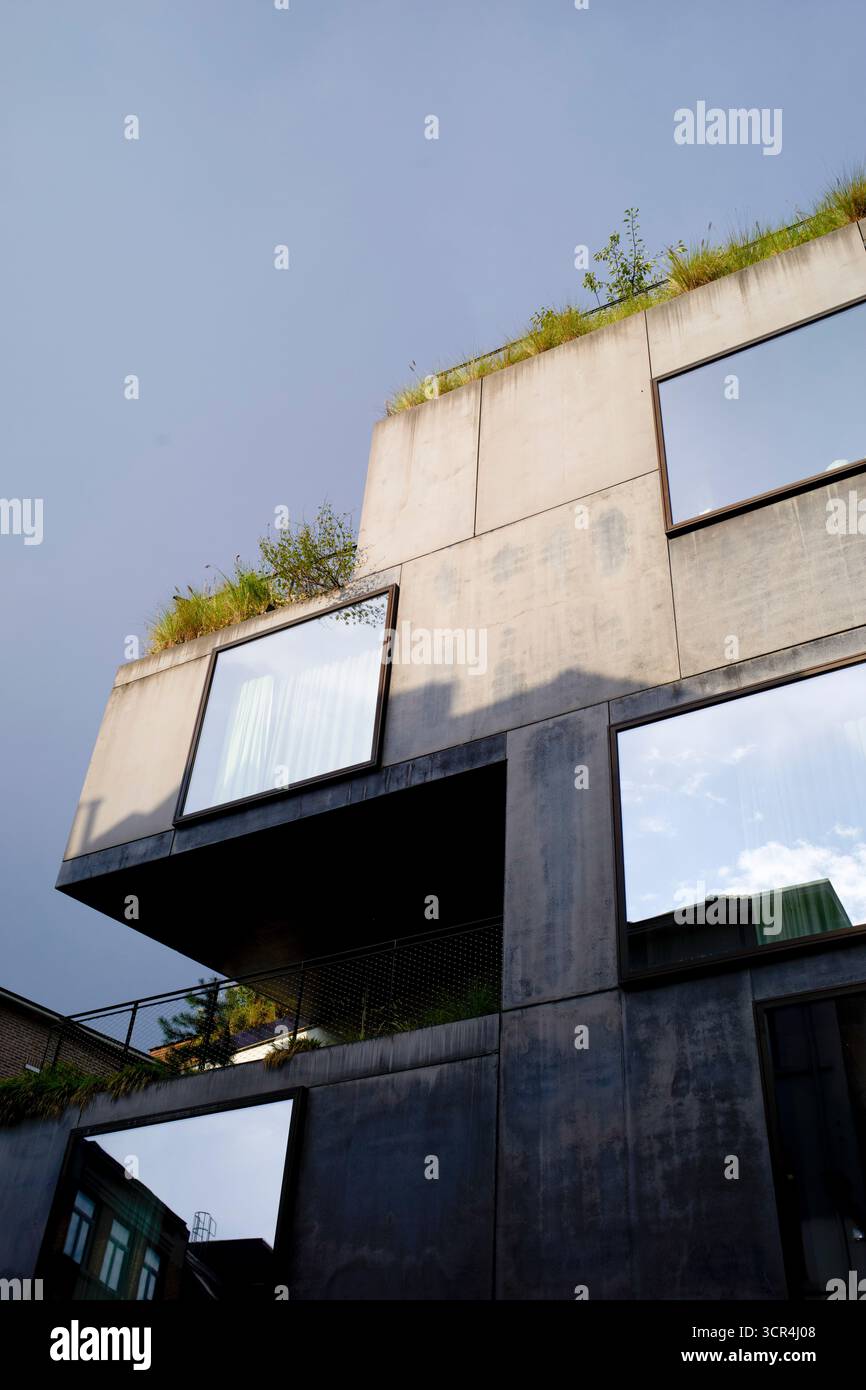 Moderno edificio in cemento con grandi finestre riflettenti e vegetazione sul tetto sotto un cielo blu. Bruxelles, Belgio Foto Stock