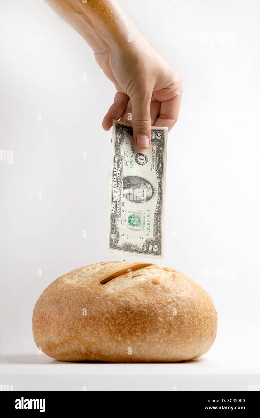 Una pagnotta di pane con una fessura per monete. La mano di un uomo inserisce una banconota da due dollari, una metafora per risparmiare o l'ironia del pane economico in un mondo costoso. Foto Stock