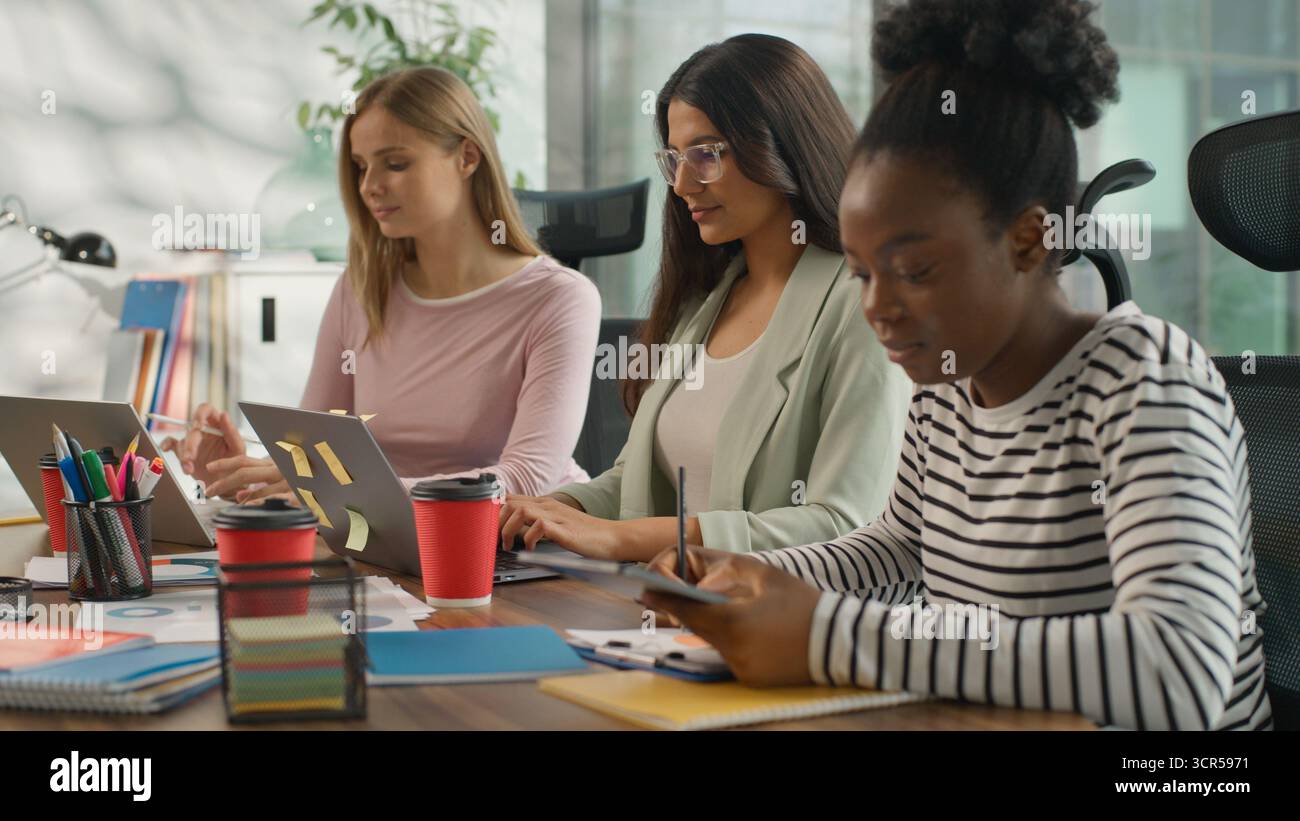 Tre donne d'affari informali multirazziali che lavorano in ufficio donne d'affari informali che lavorano con computer portatili scrivono appunti sorridenti guardando Foto Stock