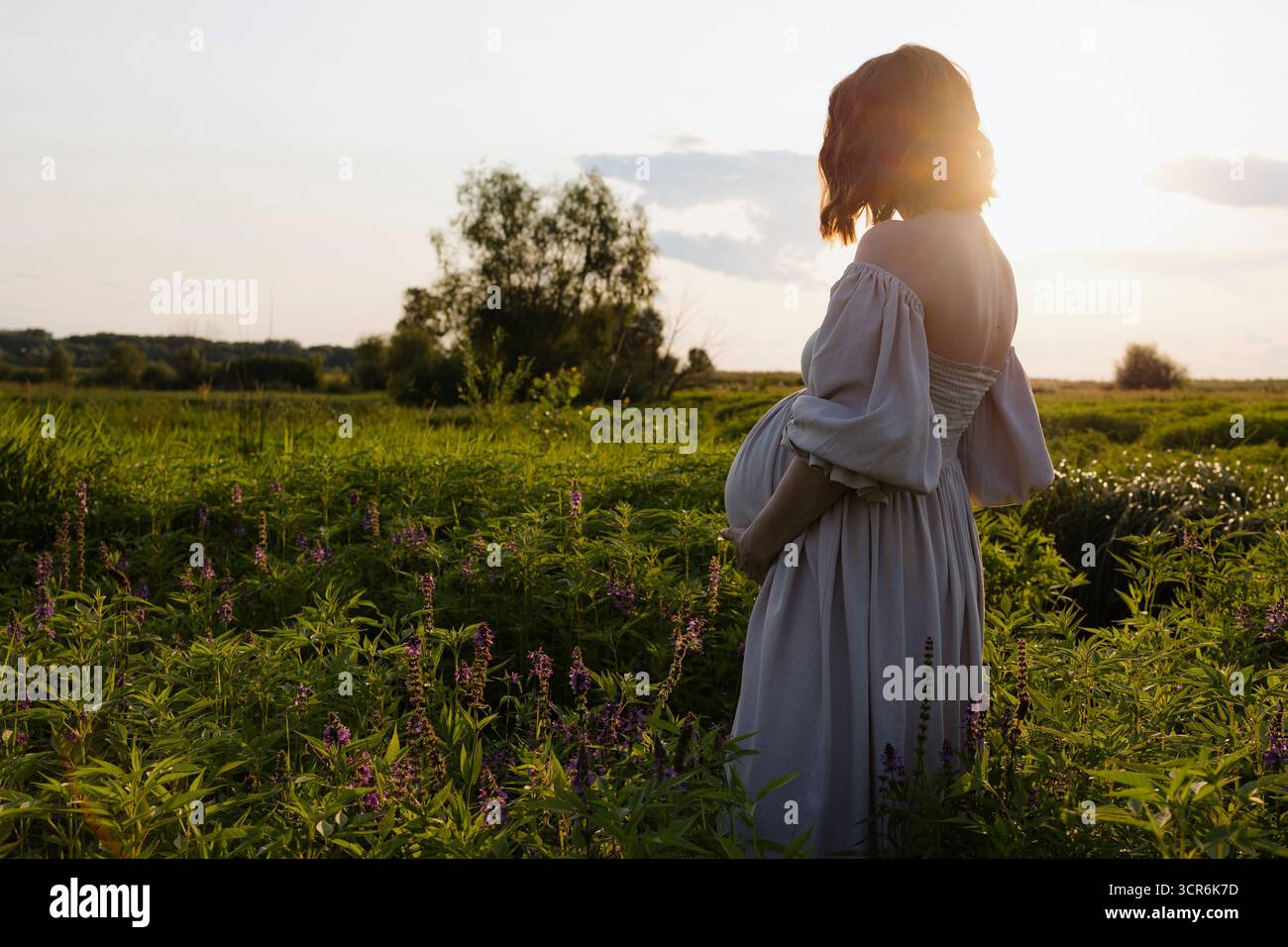 Un momento sereno cattura una donna incinta con un abito che scorre, cullando dolcemente la sua pancia mentre è circondata da una vegetazione lussureggiante mentre il sole tramonta. Foto Stock