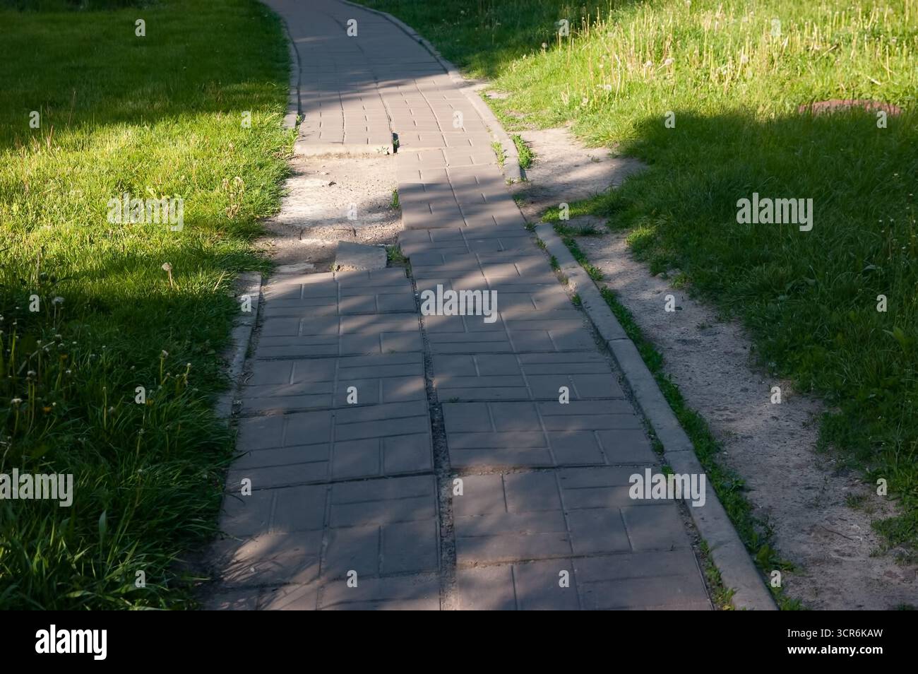Percorso piastrellato interrotto che conduce in lontananza con erba su entrambi i lati, sentiero sterrato parallelo sulla destra, ombre profonde e una zona luminosa illuminata dal sole Foto Stock