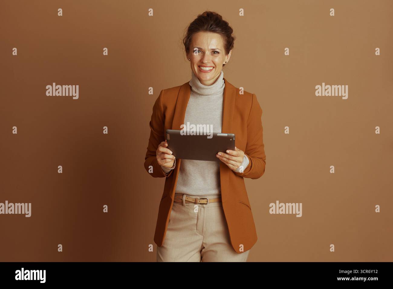 Una donna sicura di sé e sorridente, in un abbigliamento casual elegante, tiene il tablet digitale in posizione orizzontale, guardando la fotocamera. Il suo blazer marrone e la tartaruga beige Foto Stock