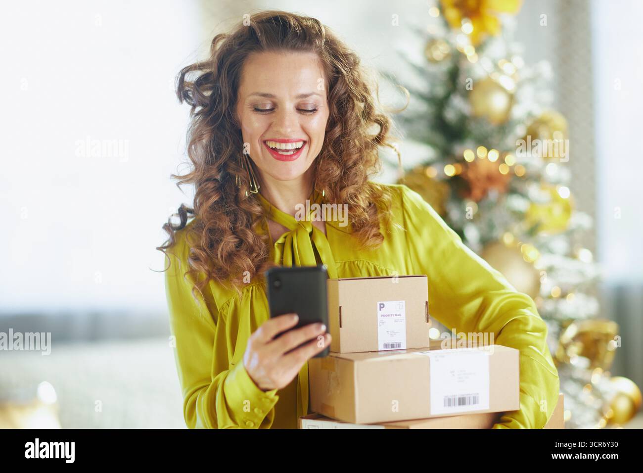 Una donna con lunghi capelli ricci ed espressione allegra può contenere smartphone e piccole scatole di cartone. Con una camicetta gialla brillante, si erge davanti a una luce soffusa Foto Stock