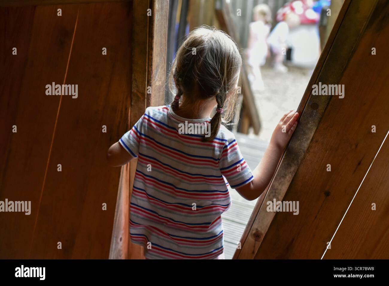 Questa immagine cattura un bambino in piedi alla soglia di una struttura in legno, guardando verso l'esterno verso uno spazio più luminoso dove giocano altri bambini. Foto Stock