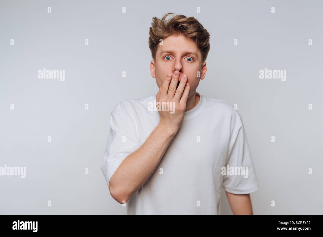 Giovane che mostra sorpresa con la mano sulla bocca in un ambiente di colore chiaro durante un momento informale Foto Stock Giovane che mostra sorpresa con la mano sulla bocca in un ambiente di colore chiaro durante un momento informale Foto Stock