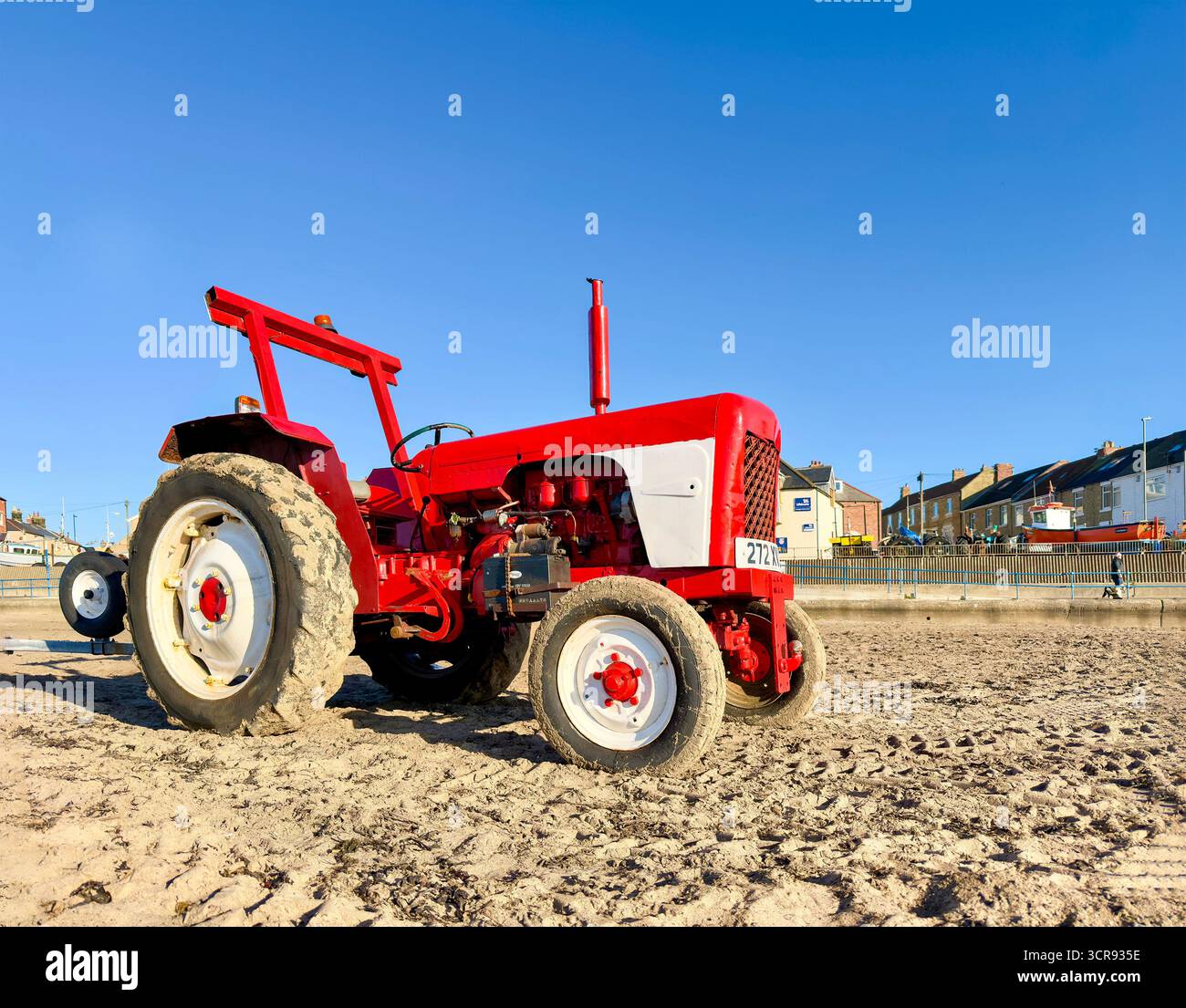 Trattore David Brown rosso d'epoca parcheggiato su una spiaggia sabbiosa con edifici costieri e un cielo azzurro luminoso sullo sfondo Foto Stock