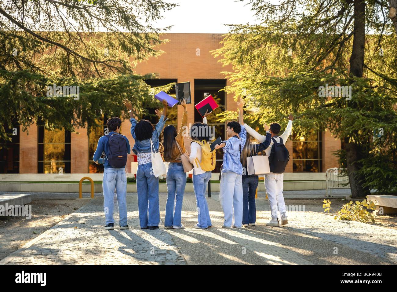 Diversi studenti visti da dietro su un percorso illuminato dal sole del campus che lancia in aria quaderni colorati per celebrare il successo accademico, l'amicizia e.. Foto Stock