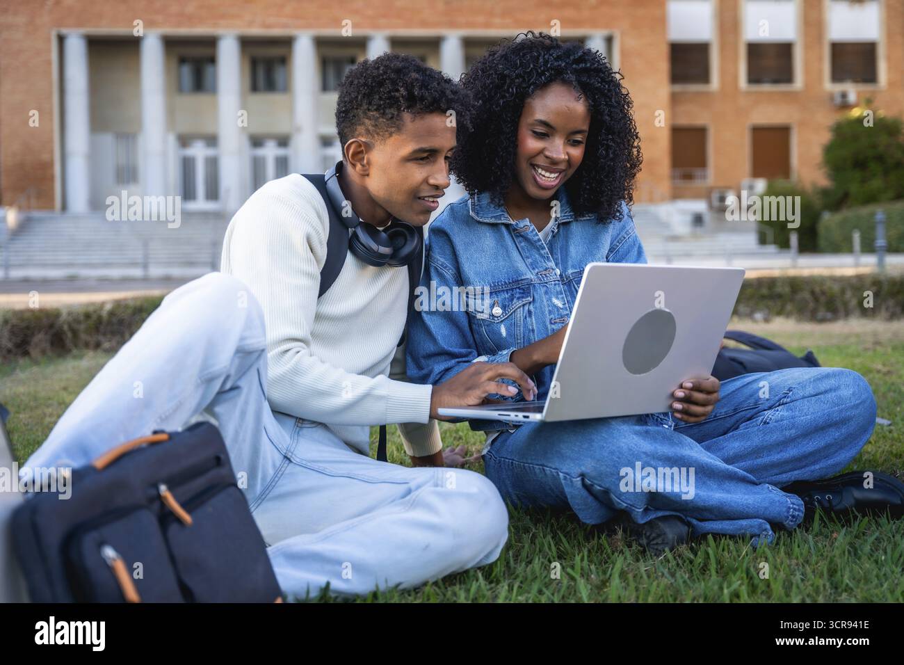 Giovani studenti neri seduti sull'erba verde, che lavorano su un notebook, discutono di progetti scolastici o contenuti di corsi online nel campus, che rappresentano l'istruzione Foto Stock