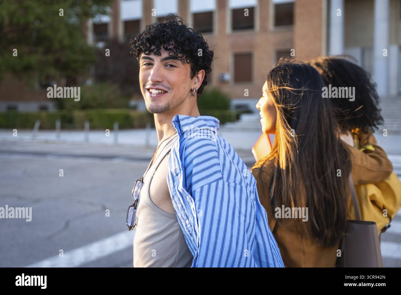 Giovane con i capelli ricci sorridente alla macchina fotografica mentre cammina con due compagne di classe su un marciapiede del campus universitario illuminato dal sole, amici informali che si divertono Foto Stock