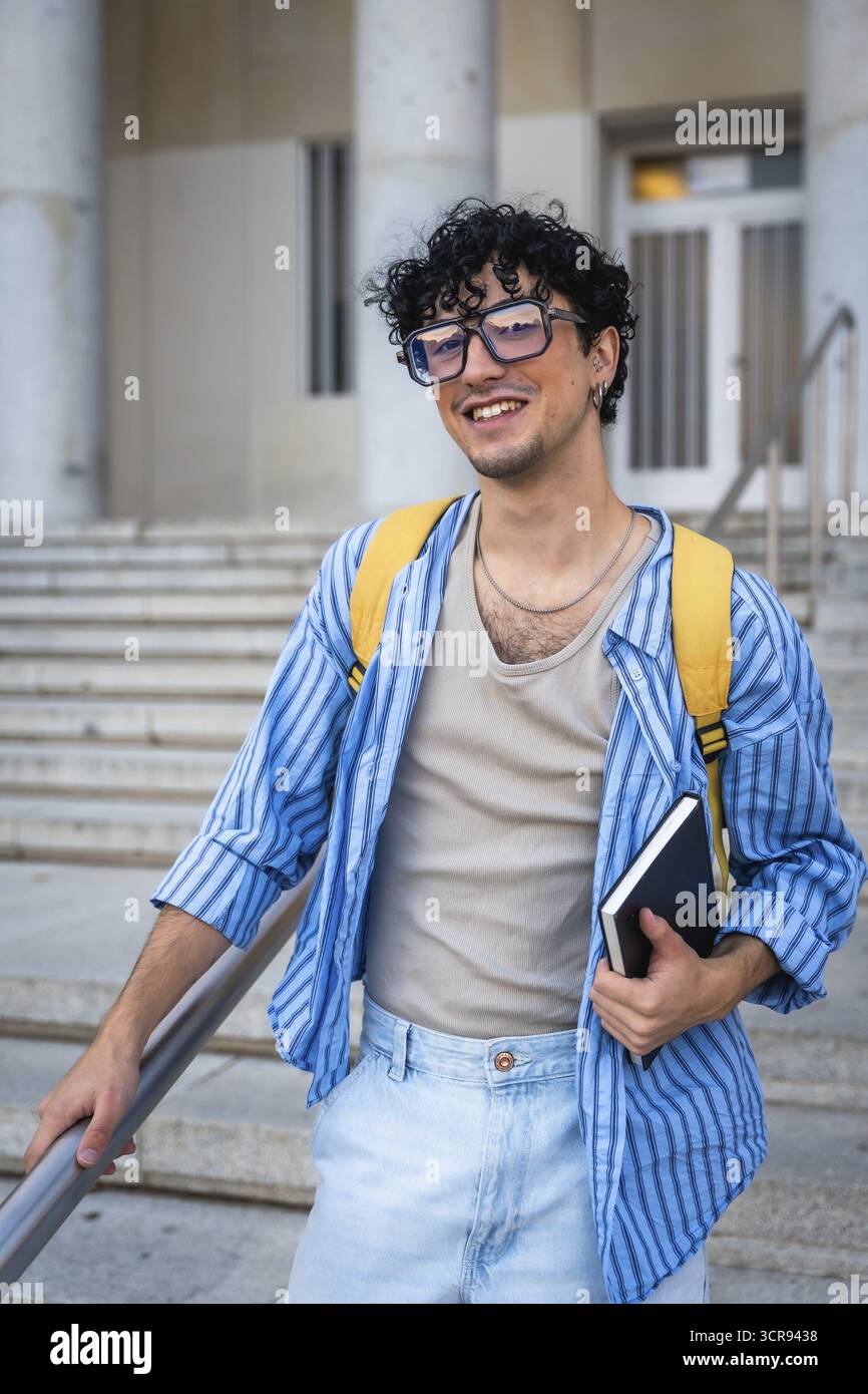 Giovane con uno zaino e un taccuino, sorridente mentre sale le scale del moderno edificio, che rappresenta l'istruzione, la vita universitaria e il futuro opp Foto Stock