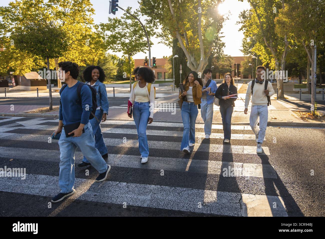 Gruppo di studenti universitari multietnici che camminano insieme lungo un attraversamento pedonale in una giornata di sole, portando libri e zaini, rappresentando l'istruzione, fr Foto Stock