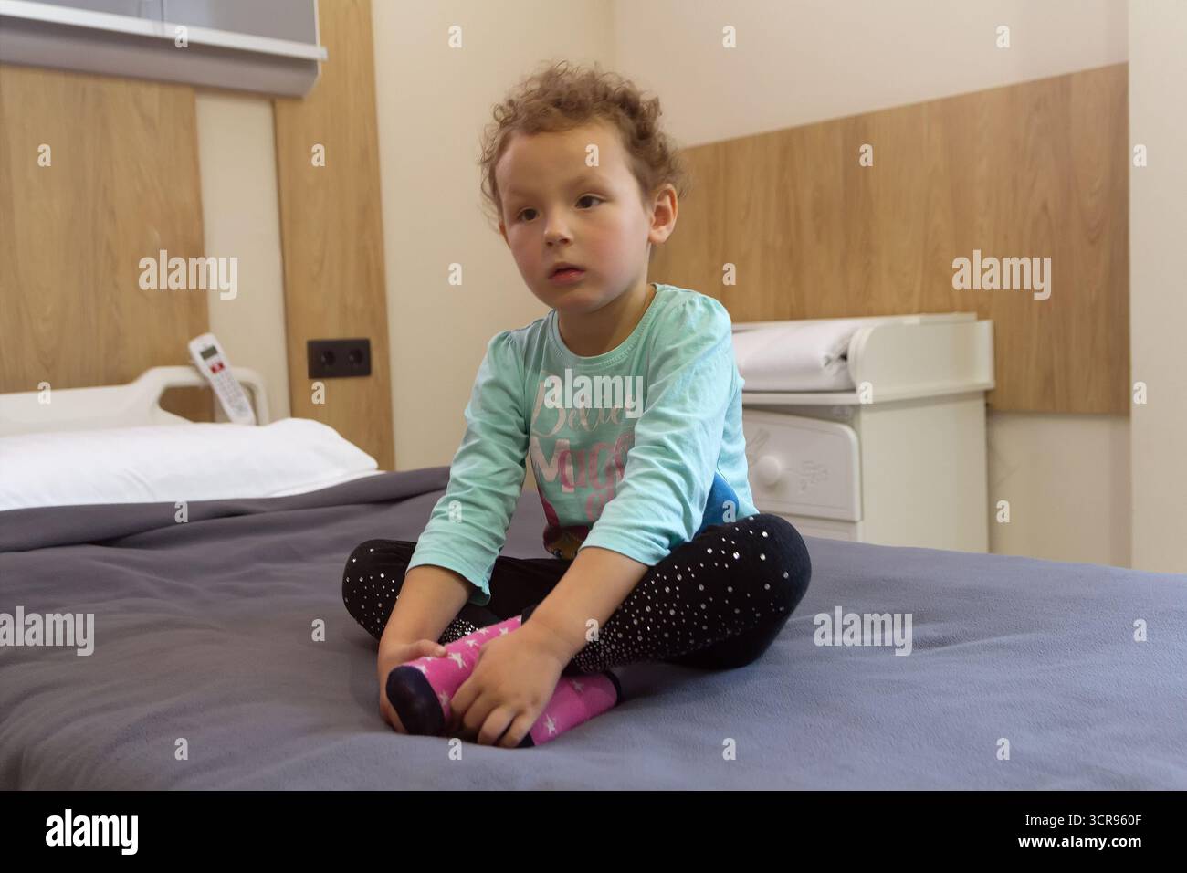 Bambino che siede sul letto d'ospedale nella stanza di visita medica, abbigliamento casual, atmosfera calma, assistenza sanitaria pediatrica. Persone Foto Stock