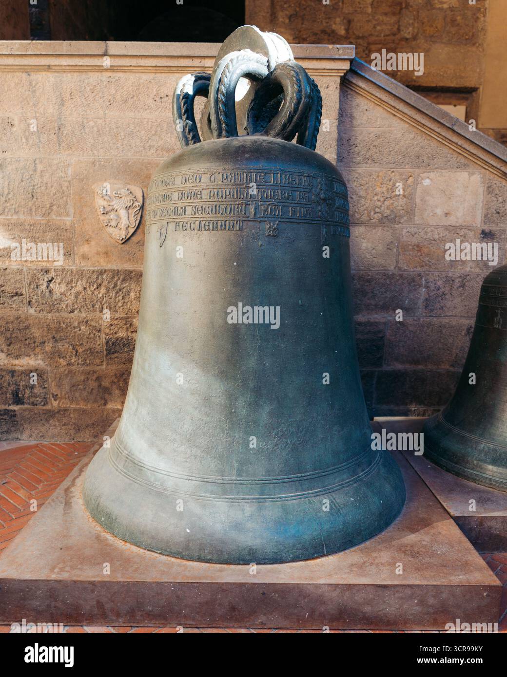 Vista di una campana di bronzo invecchiata, le sue iscrizioni intemperie che sussurrano storie di secoli passati, sullo sfondo di calde texture di pietra, San Gimignano, Toscana, Italia. Foto Stock