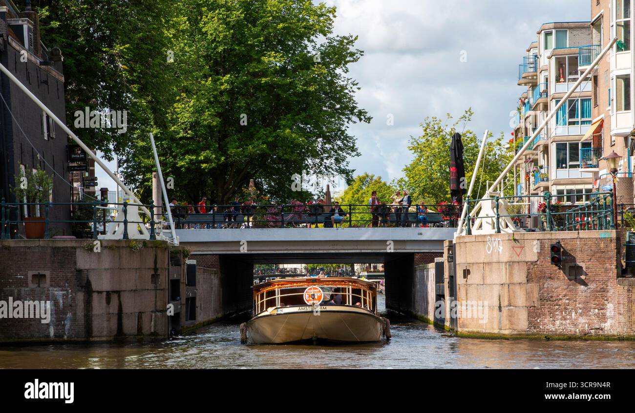 Amsterdam, Paesi Bassi - 5 agosto 2025: Un tour in barca scivola senza intoppi sotto un ponte su un canale mentre la gente gode la vista dall'alto. Foto Stock