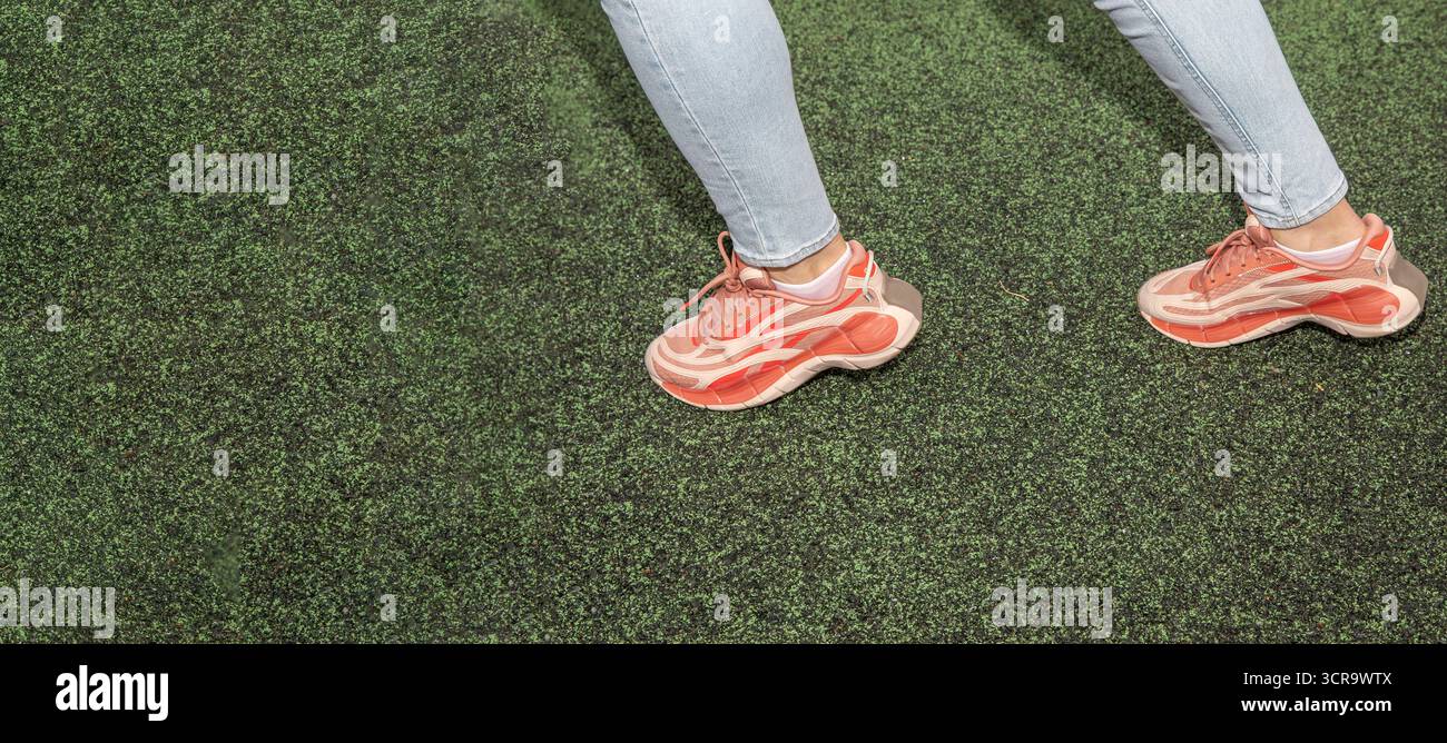 Primo piano di gambe femminili in sneakers rosse e jeans blu che camminano su un terreno sportivo verde. Concetto di stile di vita attivo, fitness, moda informale e curativo Foto Stock