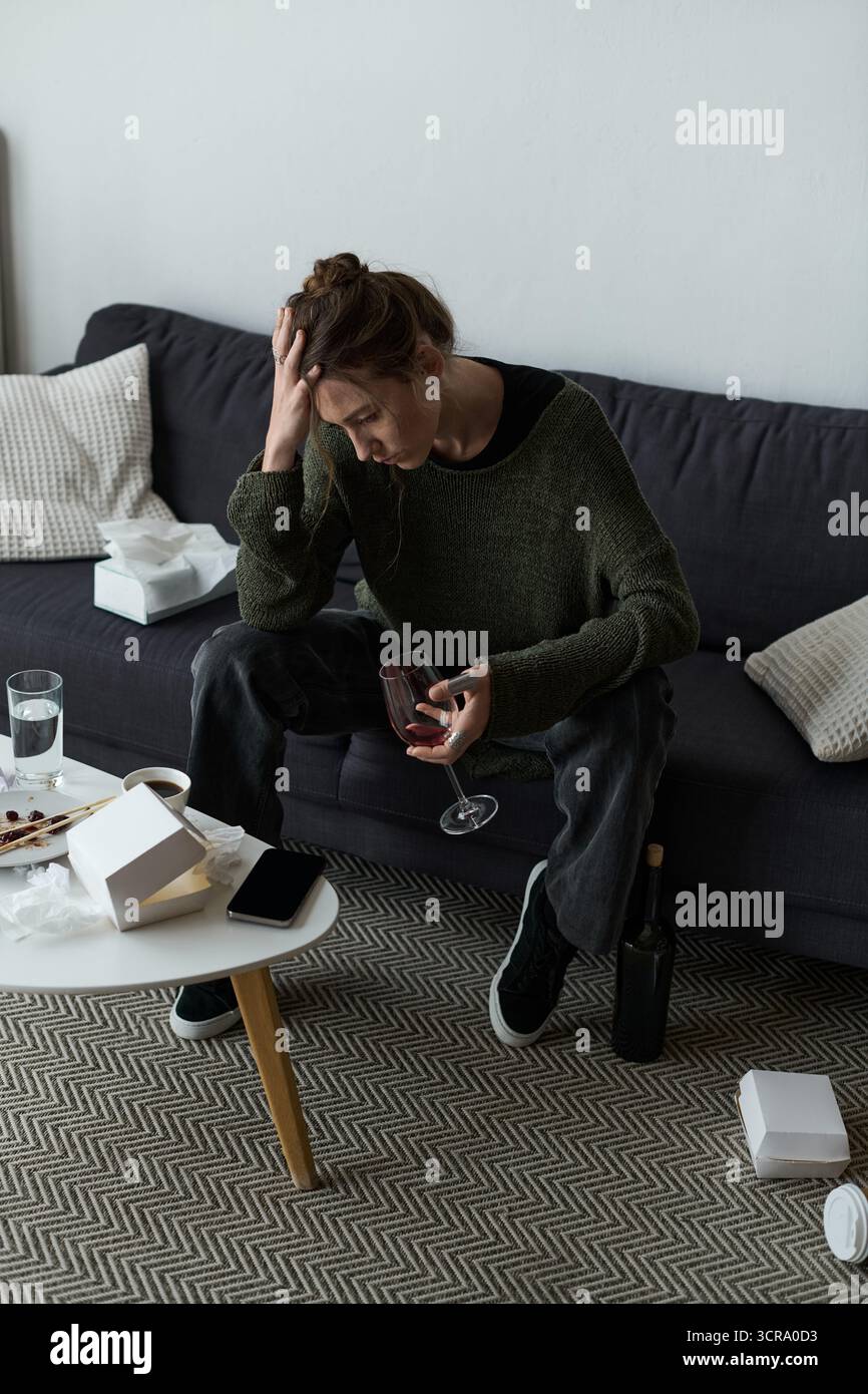 La giovane donna affronta i suoi sentimenti a casa con un drink in mano tra gli effetti personali sparsi. Foto Stock