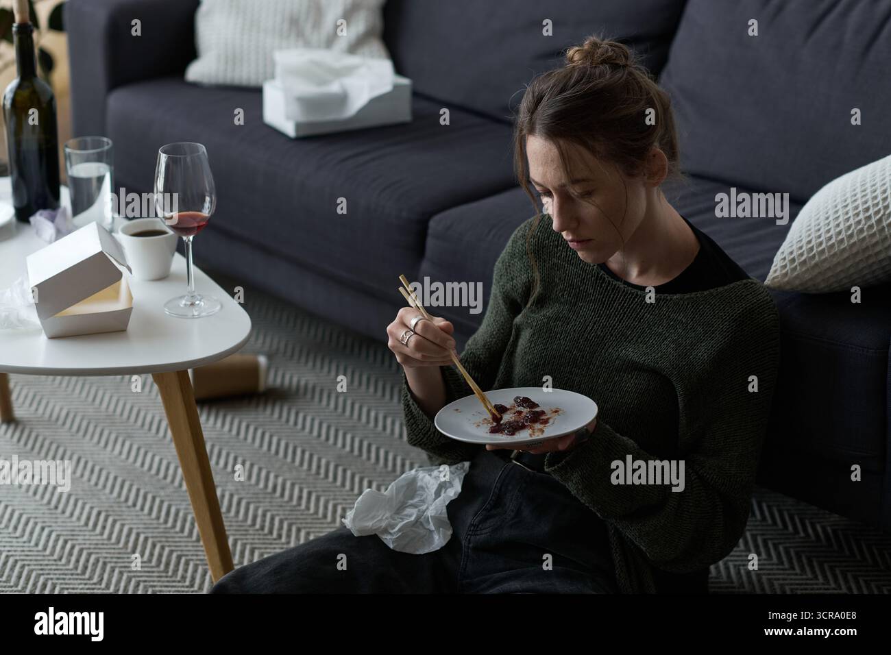 La giovane donna triste siede sul pavimento con il suo pasto, circondata da bicchieri vuoti e tessuti deformati. Foto Stock