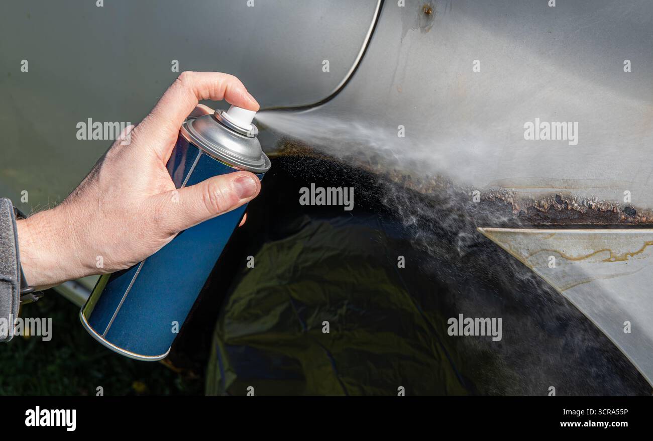 Vista ravvicinata della mano di un uomo e con una bomboletta spray per coprire la superficie di un'auto arrugginita con vernice spray bianca. Foto Stock