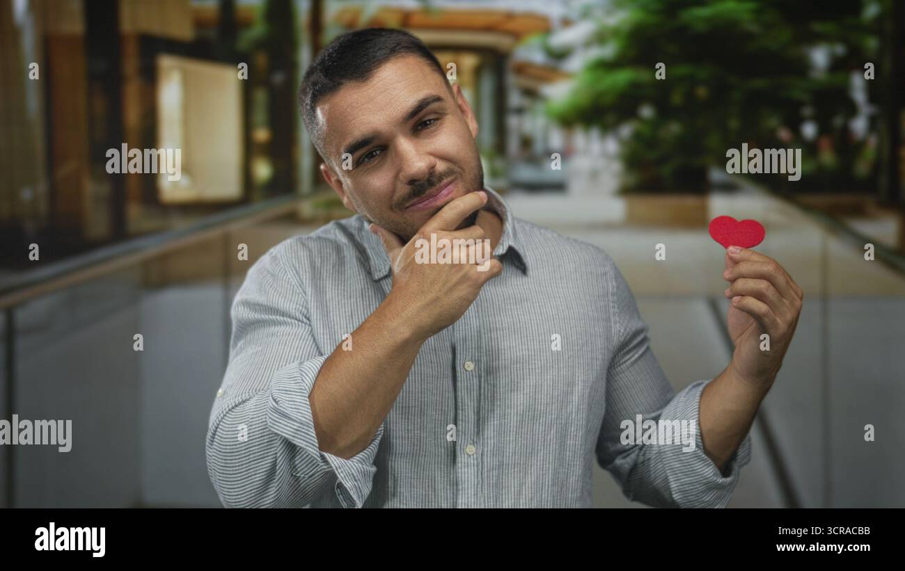 Il giovane ispanico tiene il cuore di carta rossa ritagliato con la mano sul mento per strada; riflesso romantico. Foto Stock