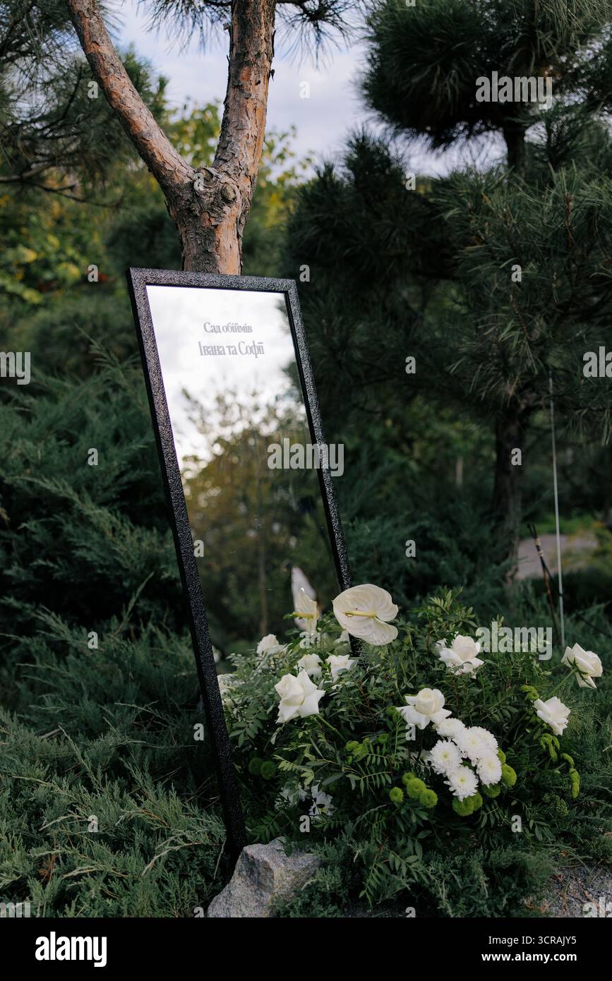 Specchio con cornice scura utilizzato come elegante cartello di benvenuto per matrimoni, collocato in giardino e decorato con composizioni floreali bianche e verdi. Traduzione da Ukr Foto Stock