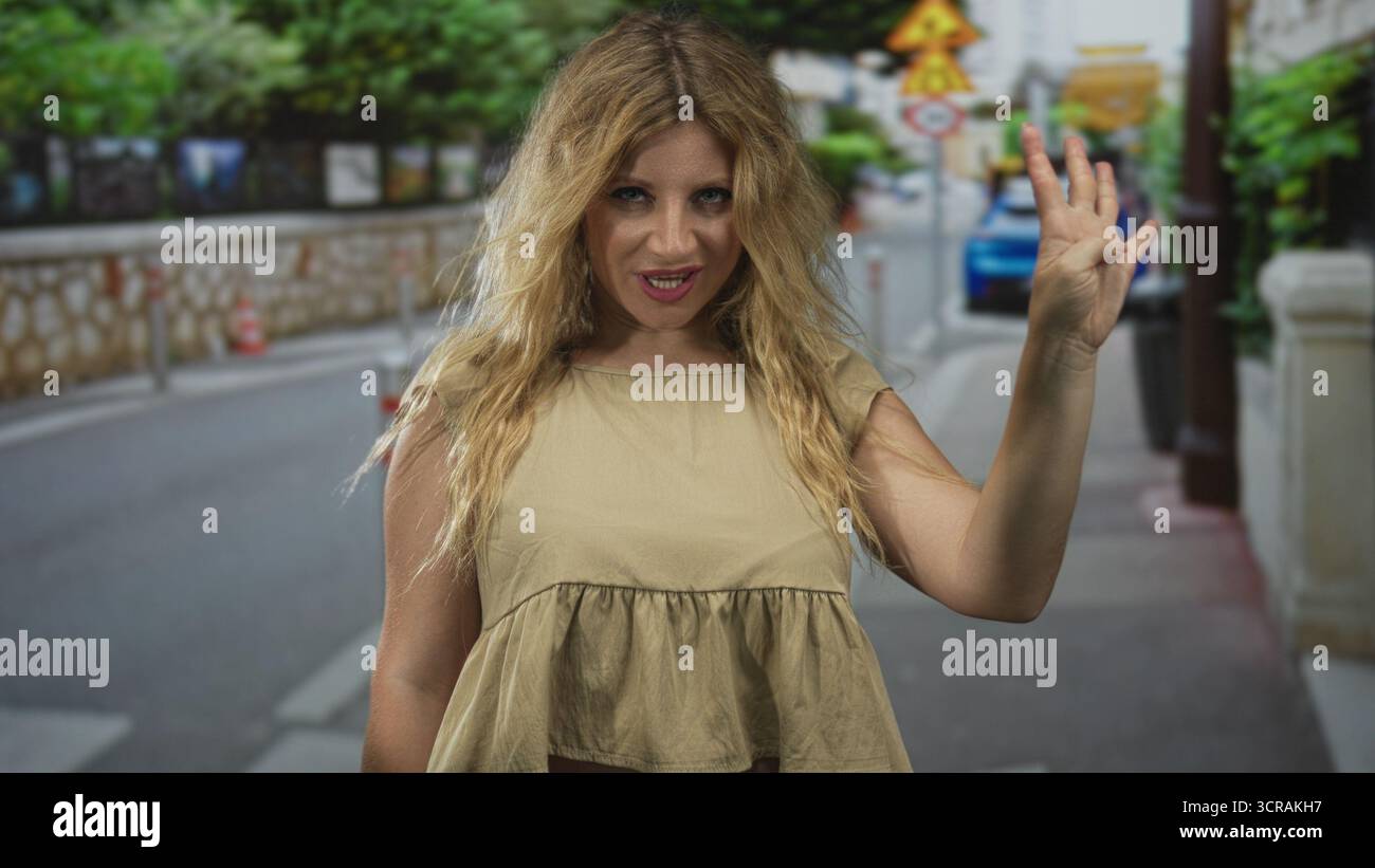 Donna che saluta la mano per strada con capelli biondi e top beige senza maniche, sorride e prepara un saluto a palma aperta alla fotocamera; un saluto amichevole. Foto Stock