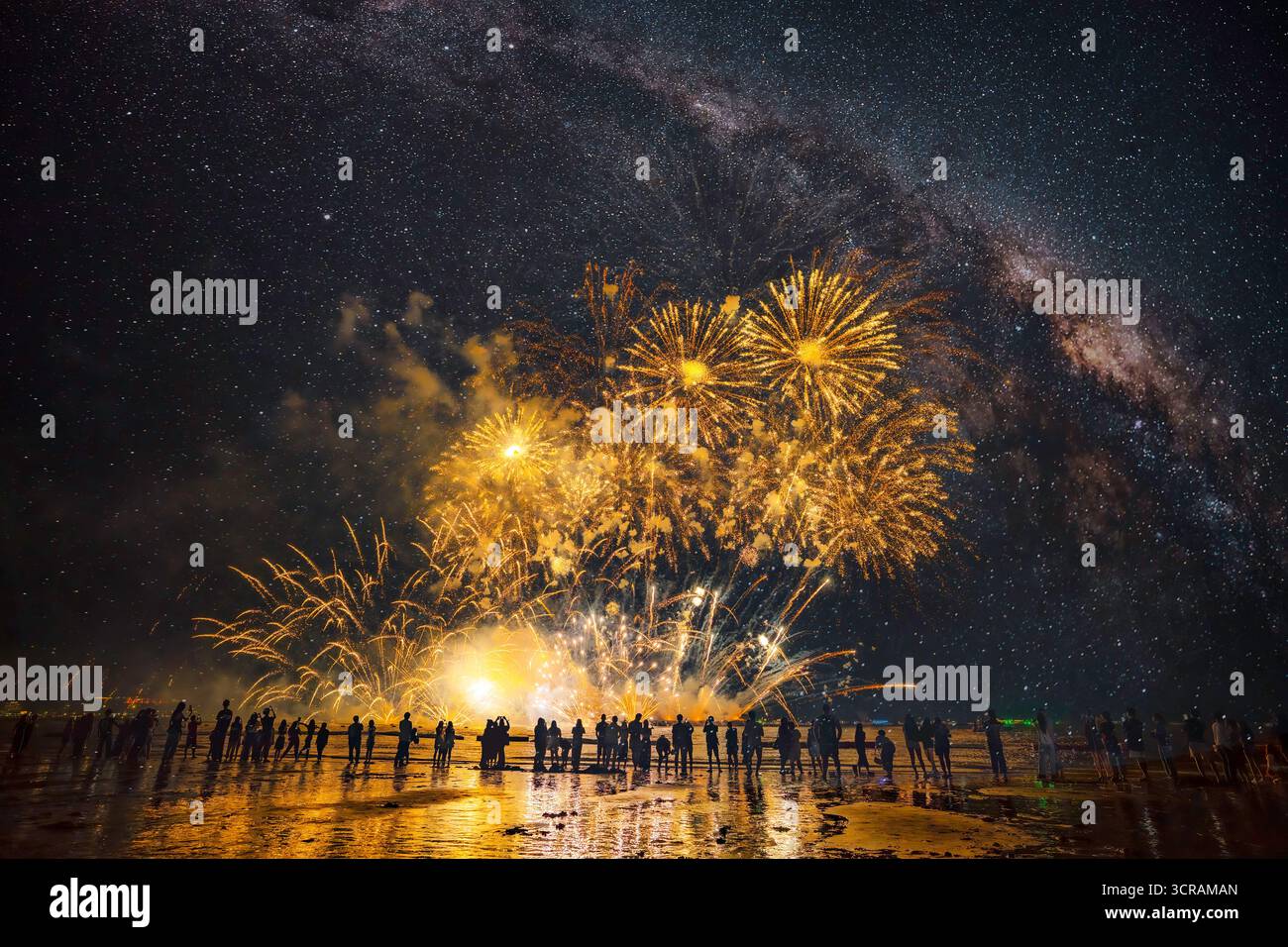 La folla sagomata guarda spettacolari fuochi d'artificio dorati che illuminano la via Lattea e il cielo notturno scuro sull'acqua, celebrando un evento. Foto Stock