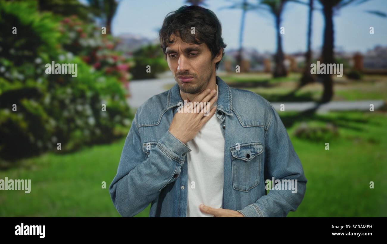 L'uomo tocca il collo con la mano mentre cammina su un sentiero lastricato nel verde del parco circondato da arbusti in fiore; fastidio alla gola. Foto Stock