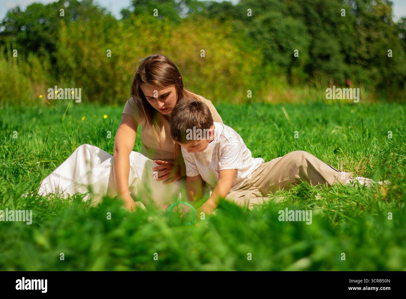 Madre che aiuta suo figlio piccolo a leggere un libro su auto e trasformatori che trascorrono del tempo sulla natura seduto sull'erba il giorno d'estate. Foto Stock