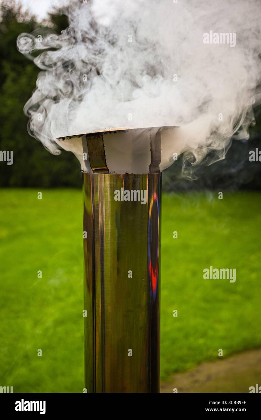 Fumo proveniente da un tubo in acciaio inossidabile lucido con erba verde sullo sfondo. Foto Stock