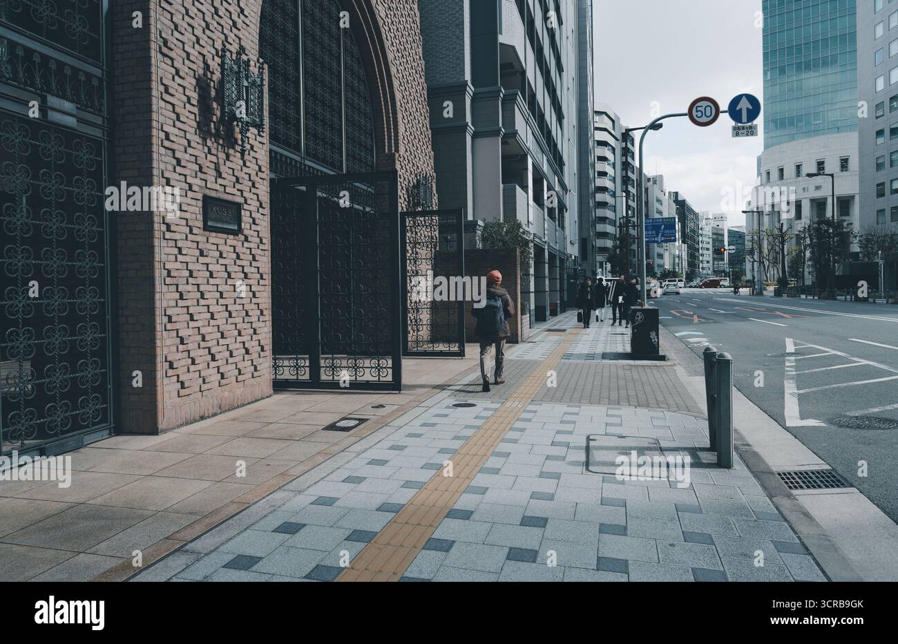Un escursionista passeggia davanti a cancelli di ferro ornati e una facciata in mattoni, dirigendosi verso una strada tranquilla fiancheggiata da alti edifici moderni a Osaka Foto Stock