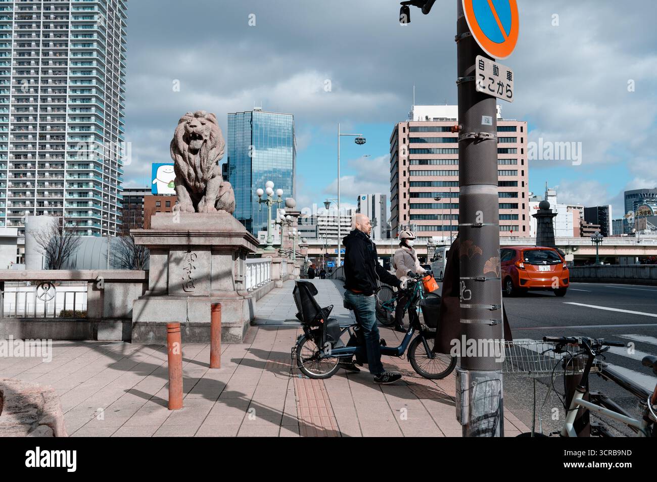 I ciclisti si fermano accanto alla statua del leone sul ponte di Naniwa, incorniciata dal moderno skyline di Osaka Foto Stock