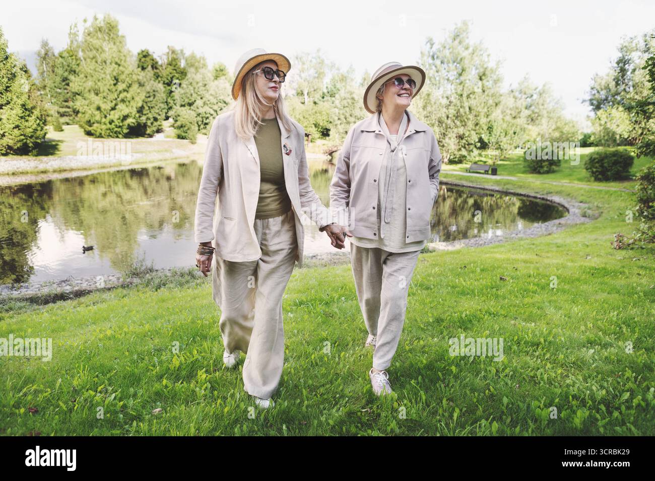 Donne anziane che camminano all'aperto con eleganti abiti in lino beige, giacche casual, pantaloni a gamba larga, cappelli e occhiali da sole, ritratti a tutta lunghezza Foto Stock