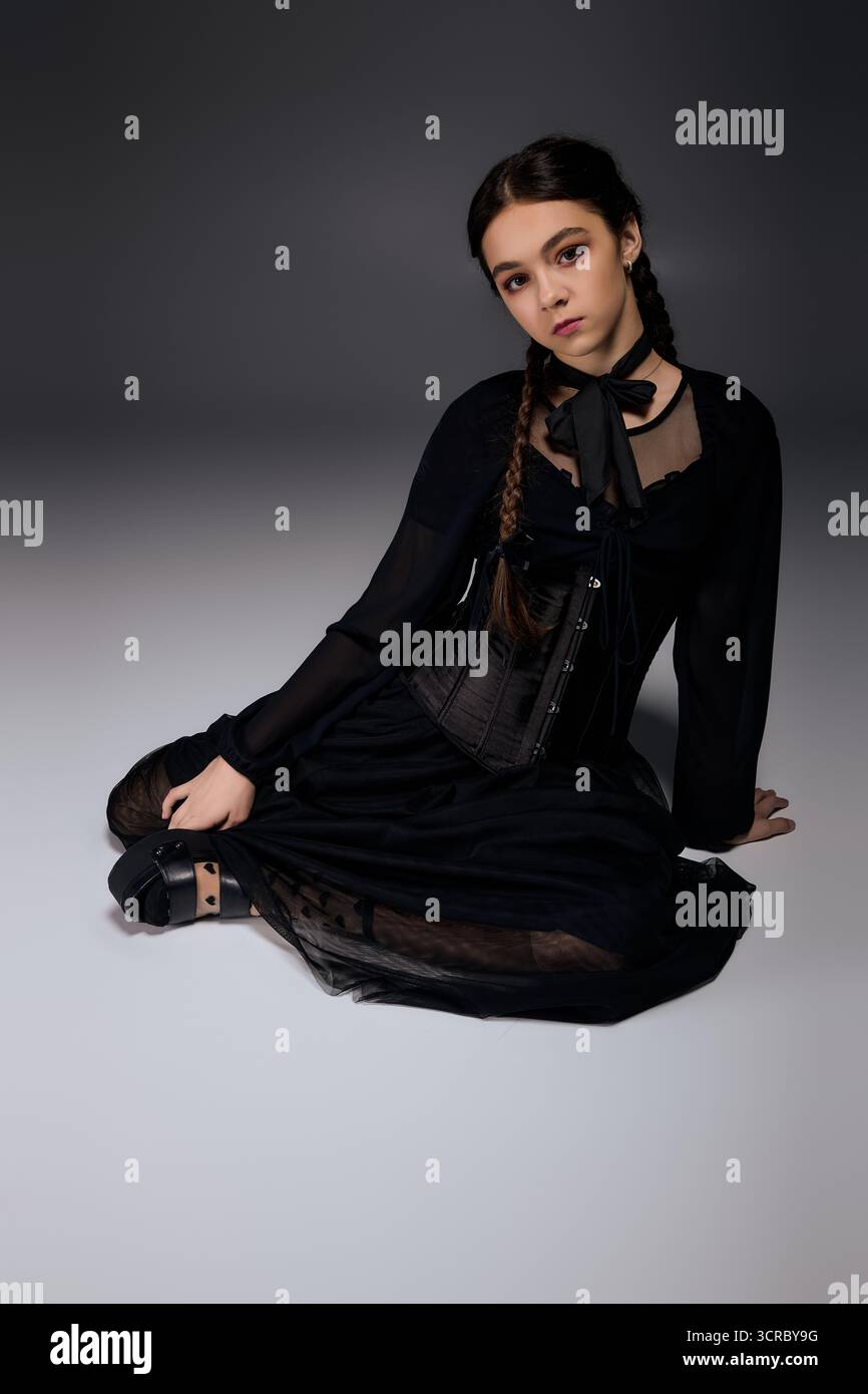 Una ragazza con trecce si siede in un abito gotico del mercoledì, catturando perfettamente lo spirito di Halloween. Foto Stock
