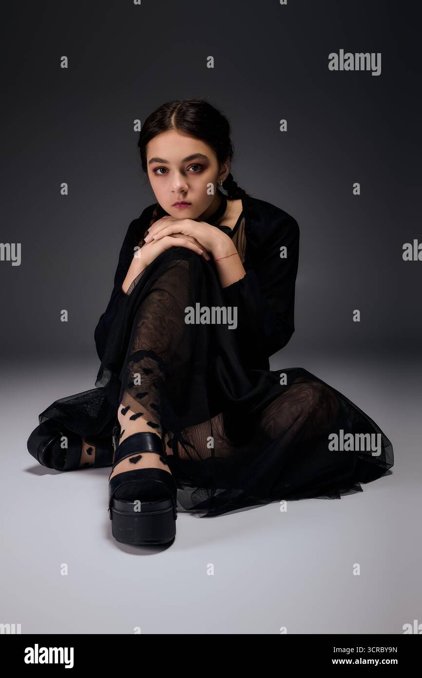 Una ragazza con trecce si siede graziosamente con un abito scuro del mercoledì, perfetto per le feste di Halloween. Foto Stock