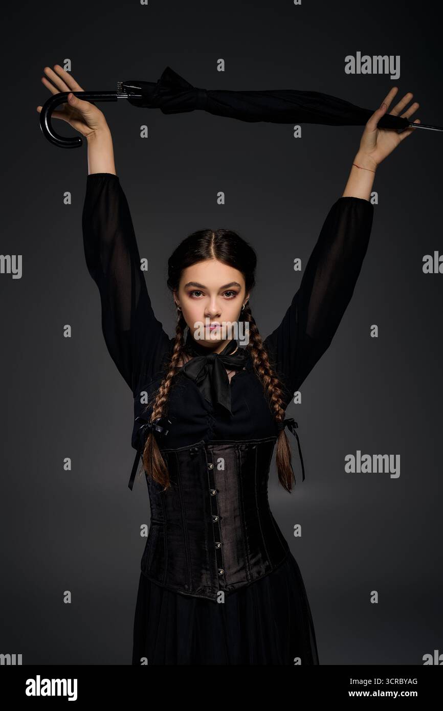 Una ragazza con trecce indossa un vestito del mercoledì e tiene in mano un ombrello nero, incarnando le vibrazioni di Halloween. Foto Stock