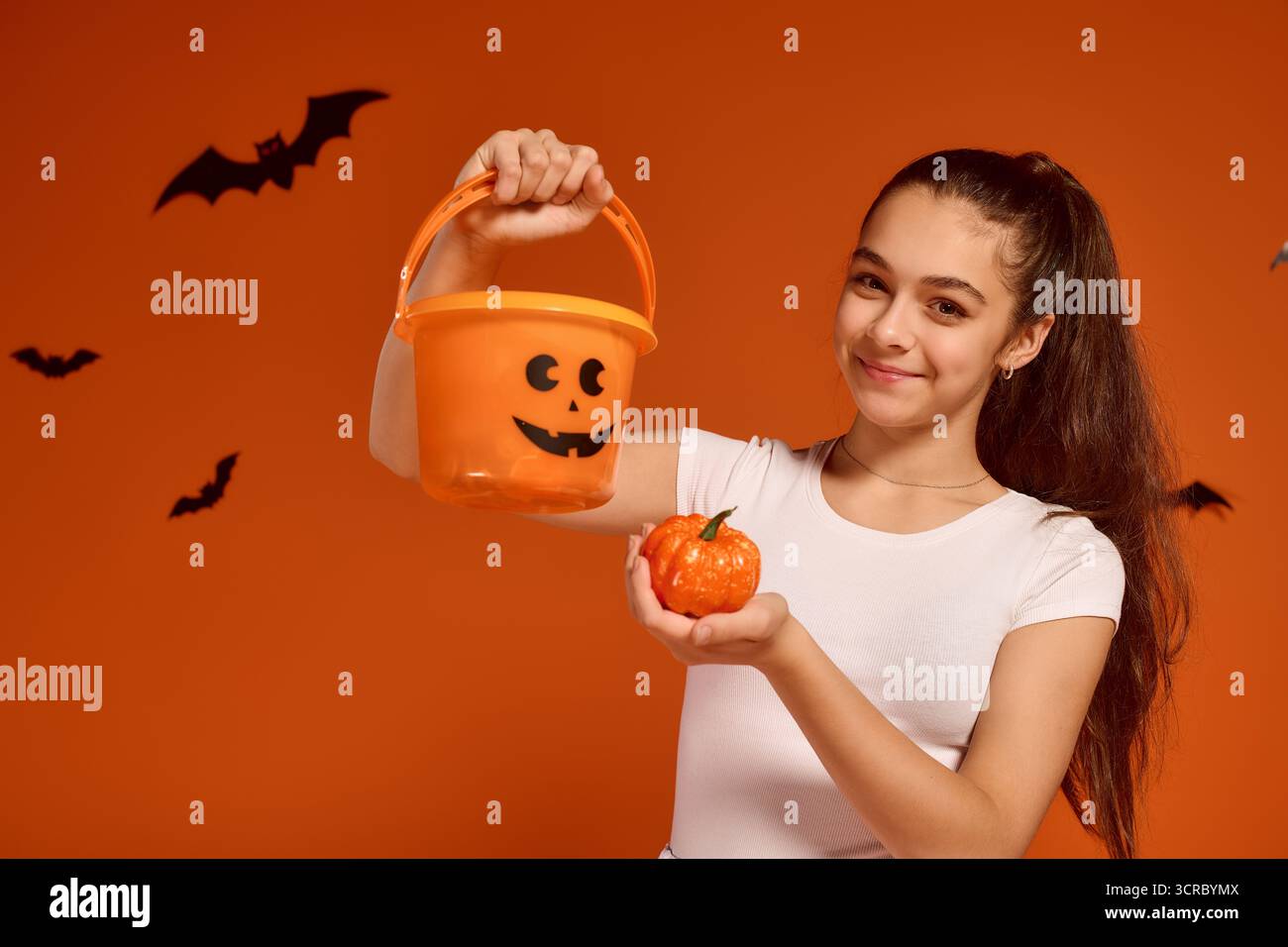 La ragazza esprime gioia per Halloween tenendo in mano una zucca e un secchio in un ambiente festoso. Foto Stock