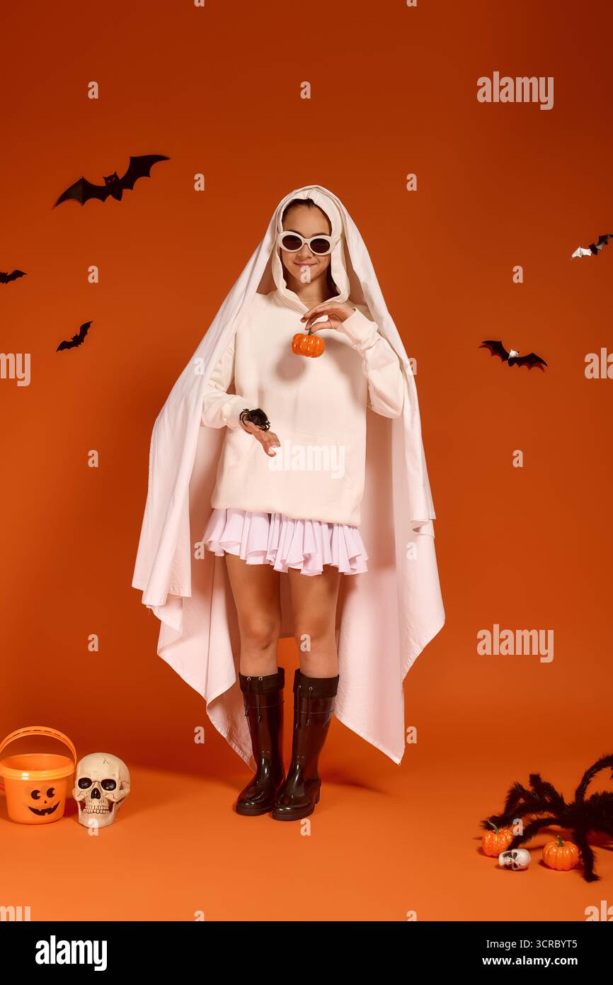 Una ragazza gioiosa vestita con un costume fantasma regala una zucca su uno sfondo arancione. Foto Stock