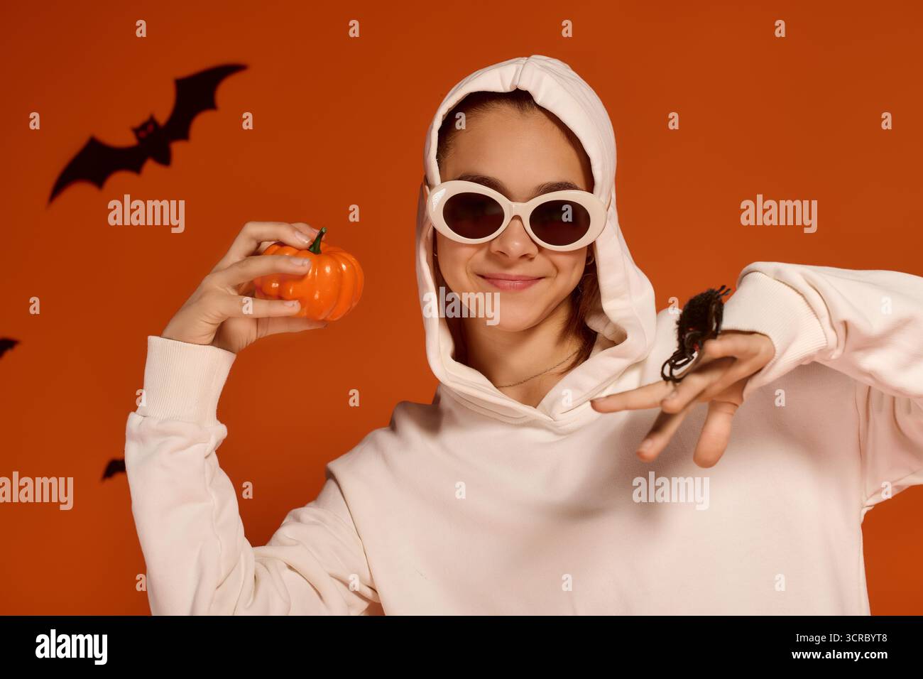 Una ragazza allegra in una felpa con cappuccio bianca tiene una piccola zucca mentre celebra Halloween con gioia. Foto Stock