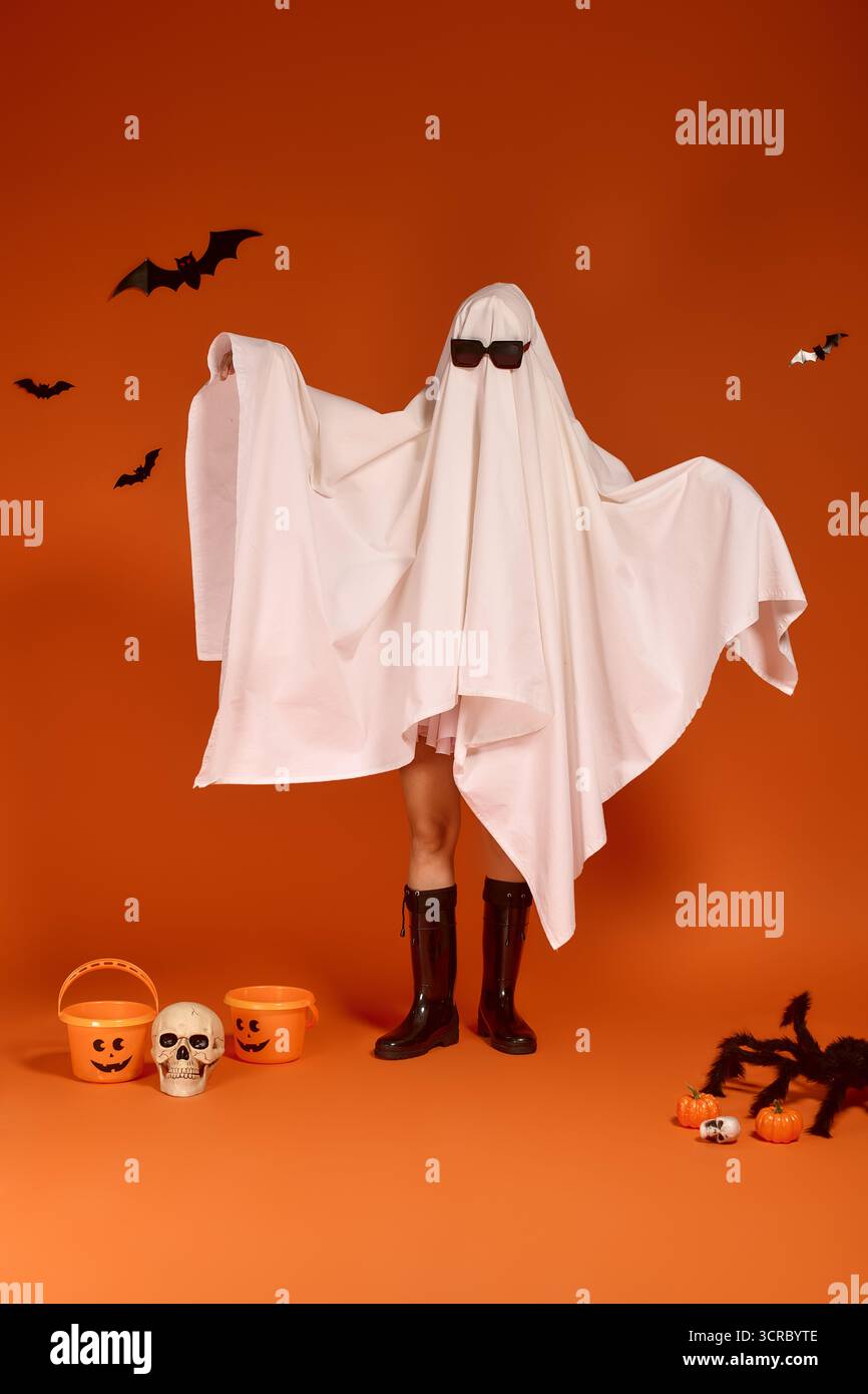 Un bambino indossa con gioia un costume fantasma e occhiali da sole su uno sfondo arancione. Foto Stock