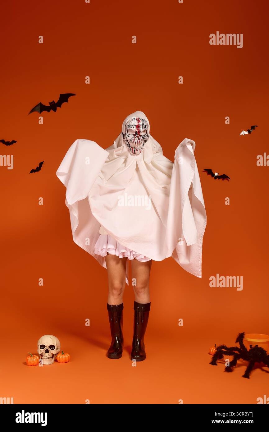 Un bambino si veste con un costume fantasma, circondato da decorazioni di Halloween e sfondo arancione. Foto Stock