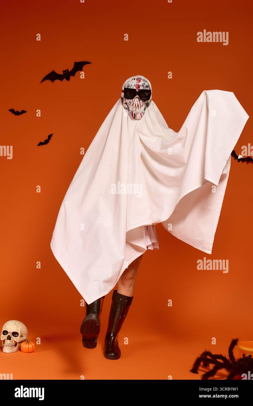 Un bambino in costume da fantasma celebra con gioia Halloween su uno sfondo arancione. Foto Stock