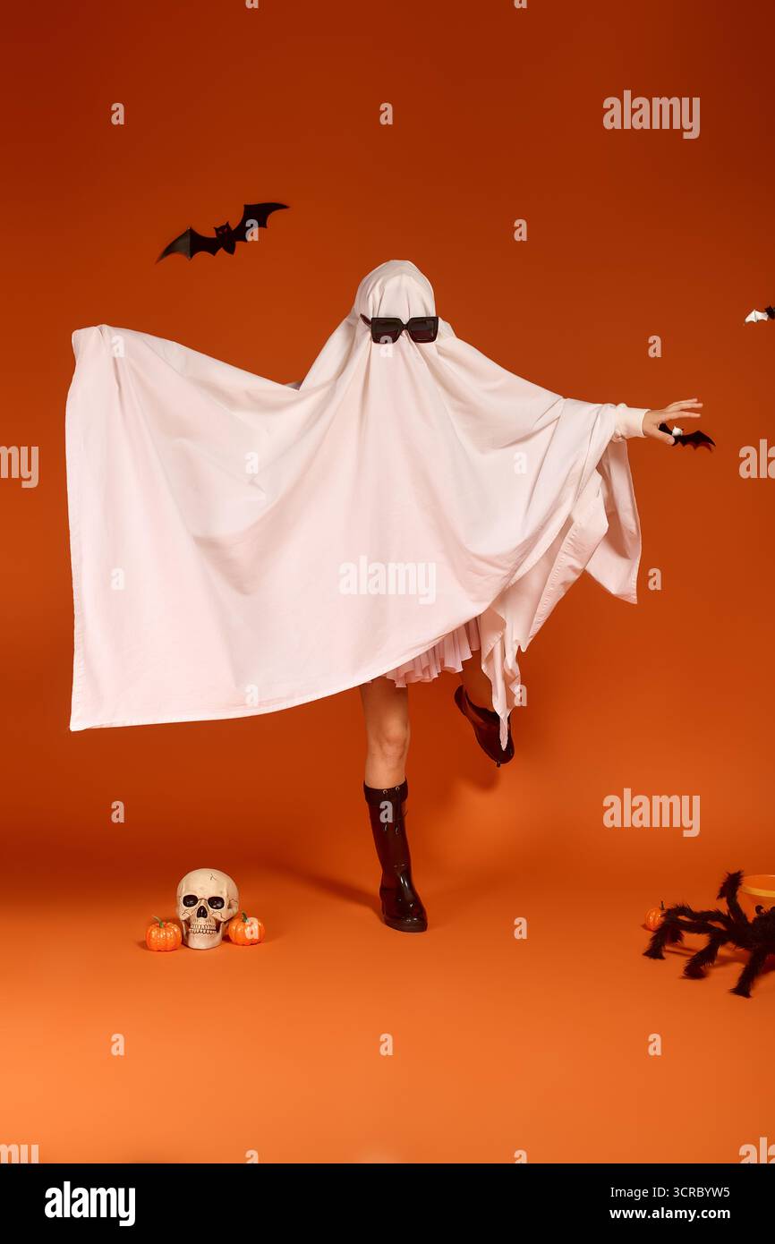 Un bambino in costume fantasma danzerà felicemente su uno sfondo arancione brillante, abbracciando Halloween. Foto Stock