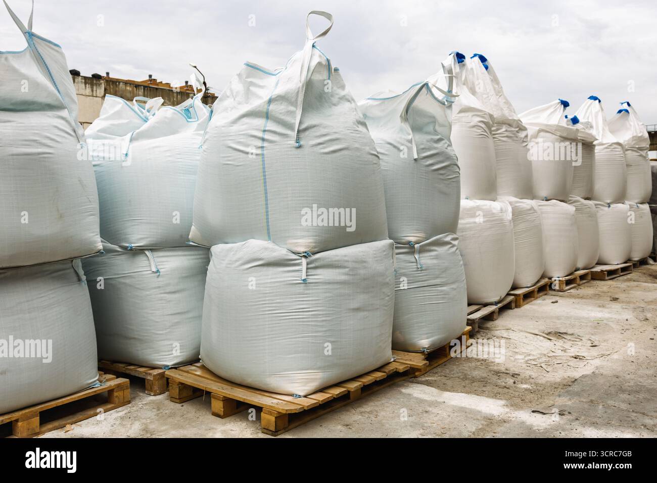 Grandi sacchetti industriali bianchi riempiti di materiali da costruzione sono impilati su pallet in legno in un magazzino all'aperto, illustrando i concetti di lo Foto Stock