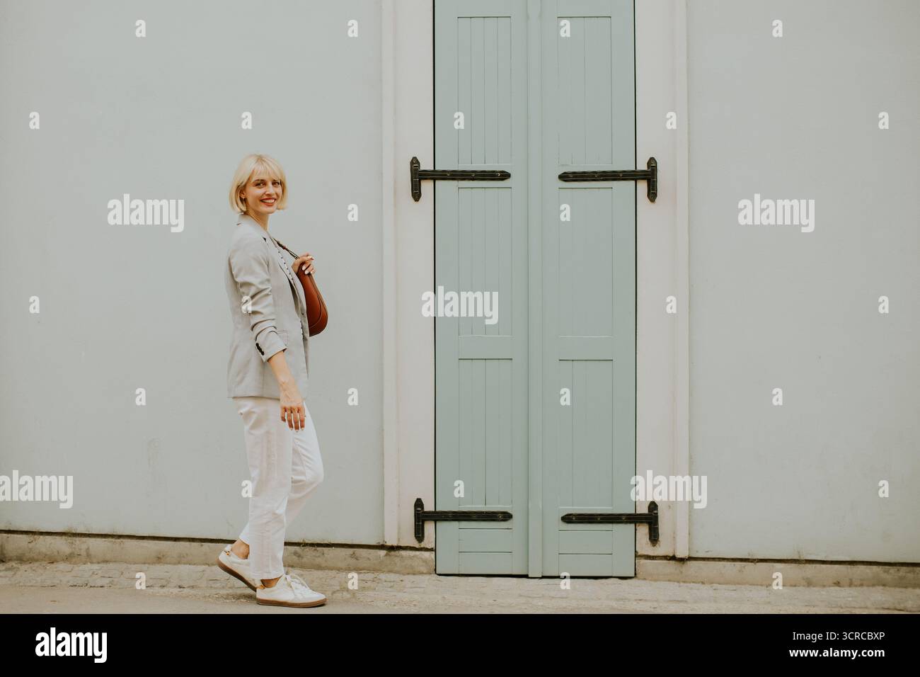 Una donna con i capelli biondi corti sorride mentre cammina accanto a una porta azzurra. È vestita con un outfit casual, godendosi una giornata di sole in un vivace urb Foto Stock
