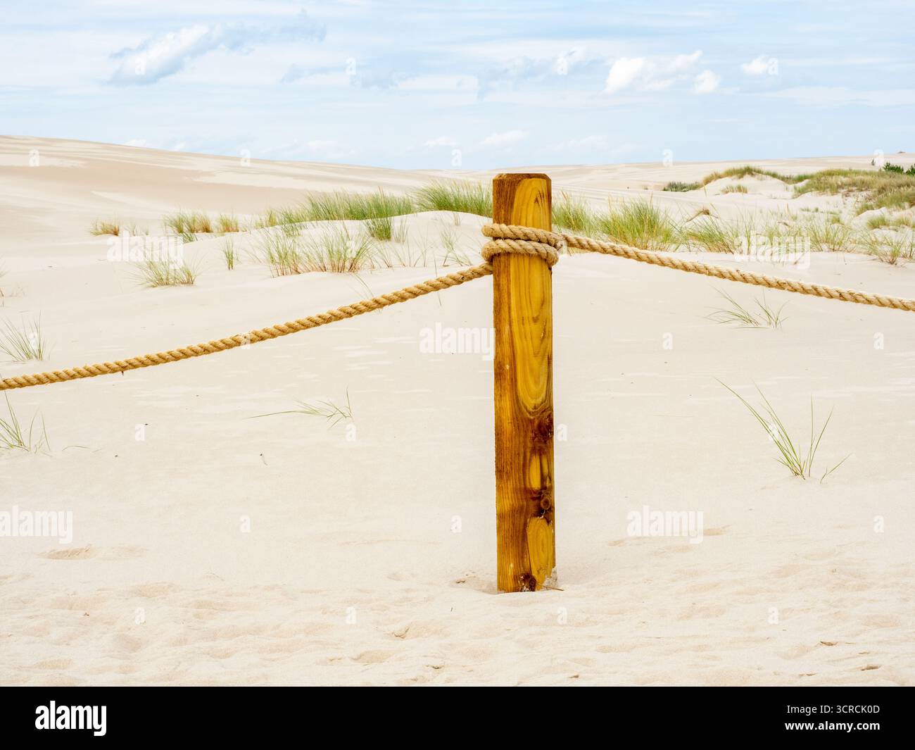 Un palo in legno resistente agli agenti atmosferici sostiene una spessa barriera di fune su dune di sabbia illuminate dal sole. Le erbe sparse punteggiano il paesaggio sotto un cielo luminoso e calmo, suggerendo Foto Stock