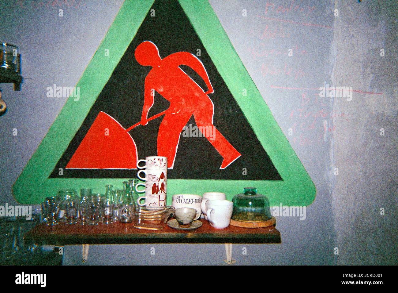 Immagine flash analogica scaduta di un cartello stradale stilizzato dipinto su una parete di una cucina accanto a uno scaffale con occhiali, creando una scena domestica stravagante. Foto Stock