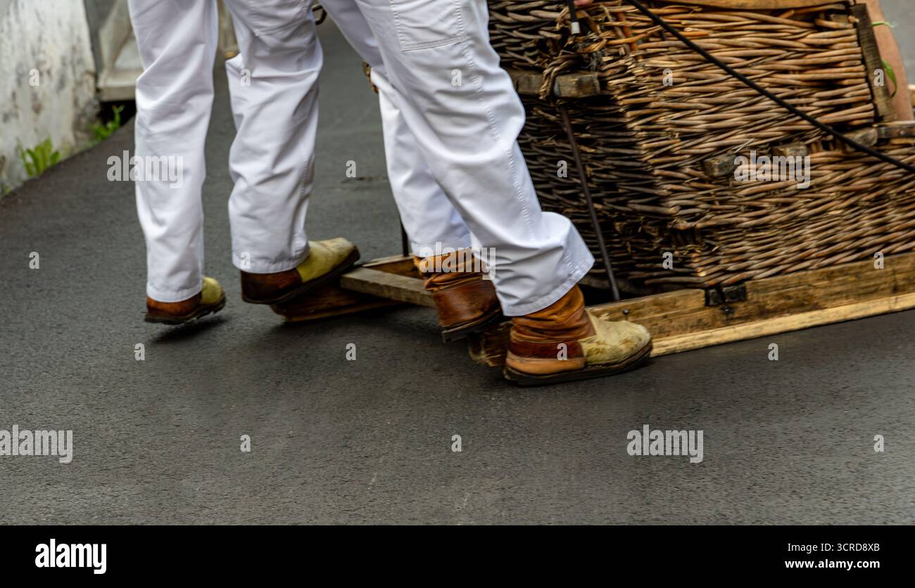 Due persone in tradizionale abbigliamento bianco manovrano una slitta di vimini su una strada liscia pavimentata vista dal retro Funchal Madeira Portogallo Foto Stock